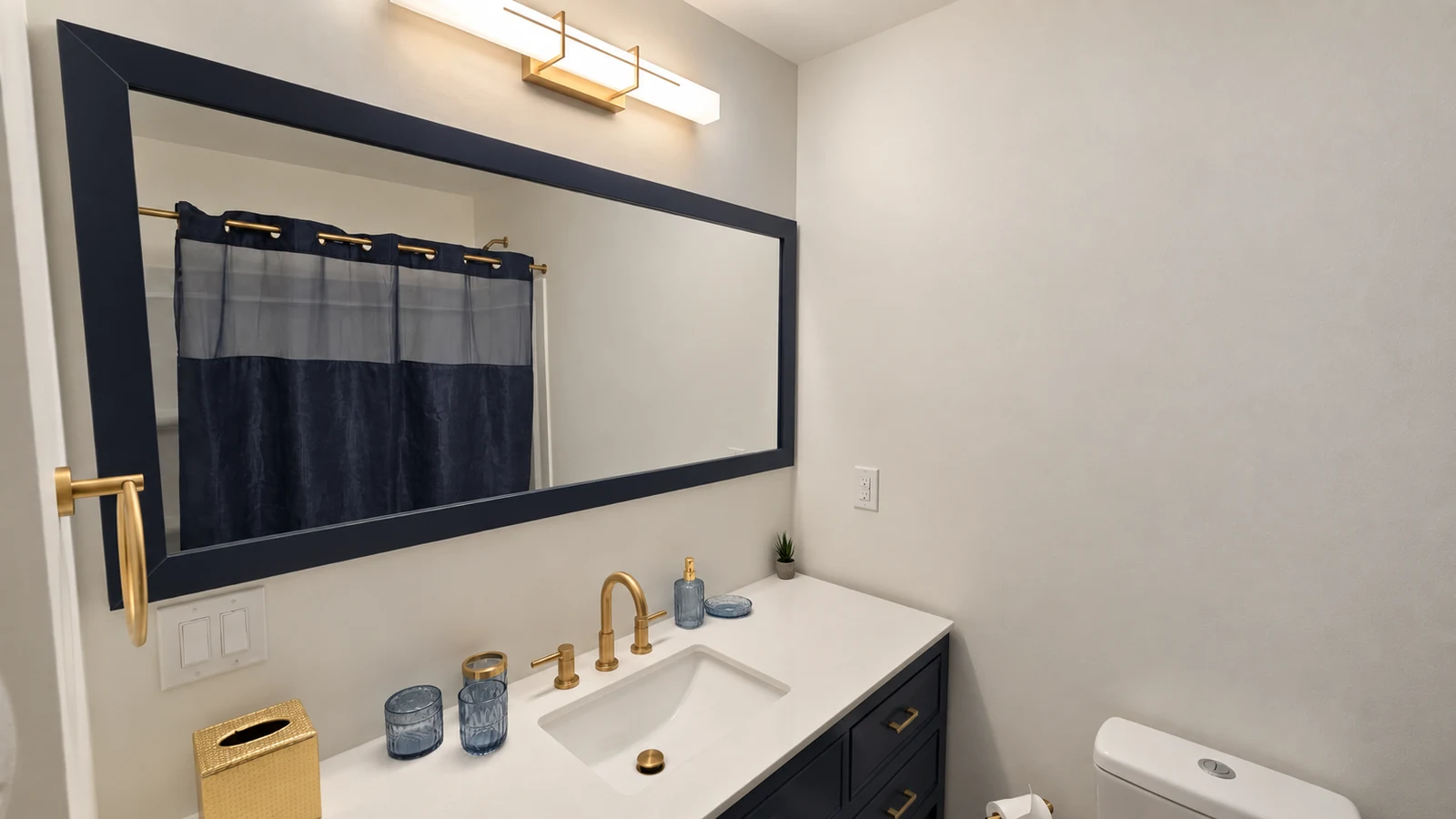 Hall bathroom vanity with gold faucet, navy mirror, and designer accessories